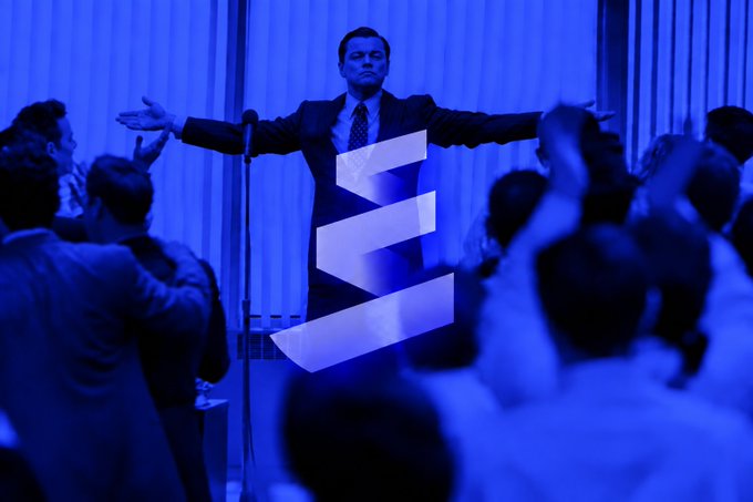 A man in a suit stands on a stage with arms outstretched, bathed in blue light with a large 'S' logo overlay, in front of an applauding audience.