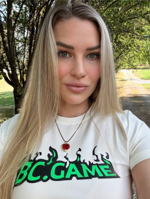 A young woman with long blonde hair poses outdoors, wearing a white t-shirt featuring "BC.GAME" branding in bold green and black text with flame graphics, along with a gold heart pendant necklace. The image ties to the post about BC Engine's reward distributions by displaying BC.Game apparel, visually reinforcing the crypto gaming platform's branding alongside the announcement of $8.6M in payouts to $BC holders.