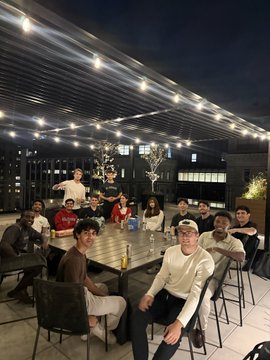 A group of smiling colleagues from Sideshift gather around a long outdoor table under string lights at night, posing together for a team photo. The image captures the camaraderie of their remote-friendly company culture, complementing the post's lighthearted jab at solo remote work while showcasing the team that builds their global distribution product.