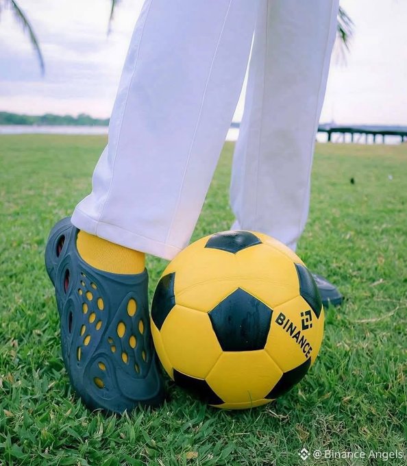 A person's foot in a teal croc rests against a yellow and black soccer ball branded with "BINANCE," standing on grass with water in the background. The image playfully nods to the "GM!" (Good Morning) post with its casual outdoor soccer scene evoking a relaxed morning vibe.