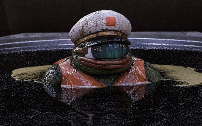 A close-up of a small, weathered toy figure resembling a sailor or military character, wearing a white hat with an orange square, green skin, and an orange life vest, sitting in dark water.