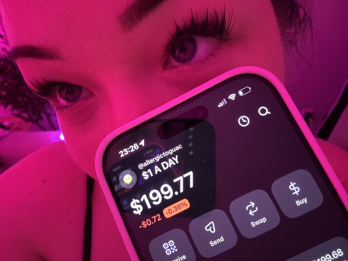 A close-up selfie of a person's eyes partially covered by a smartphone displaying a crypto/stock app showing '@aftergrctoguac' with '$199.77' down -$0.72 (-0.36%) for $1 A DAY, featuring Send, Swap, and Buy buttons.