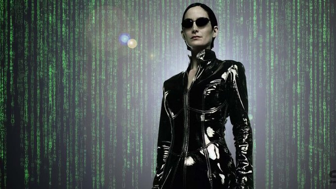 The image features a woman wearing a black leather outfit, standing against a green background with a pattern of small square lights. The woman appears to be posing for the photo, possibly as part of an advertisement or promotional material related to the brand "Polkadot Trinity".