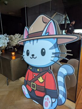 A cartoon cat mascot dressed in a red shirt and safari hat stands in a modern indoor venue, likely representing the Hyperliquid Toronto Social event mentioned in the post. The image appears to be a decorative life-sized cutout, complementing the promotional content about the Hyperliquid Summit 2026 gathering.