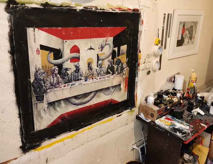 An artist's studio features a large, framed painting parodying The Last Supper with fantasy creatures, mounted on a paint-splattered wall beside a cluttered worktable and framed artwork.