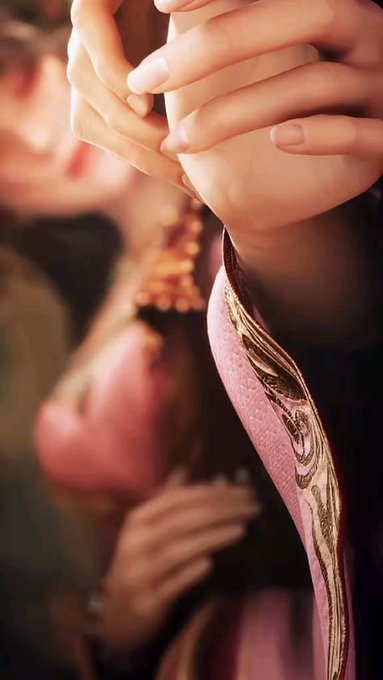 Close-up of hands delicately cupped together, with a blurred figure in pink and gold attire visible in the background, creating a warm, intimate atmosphere.