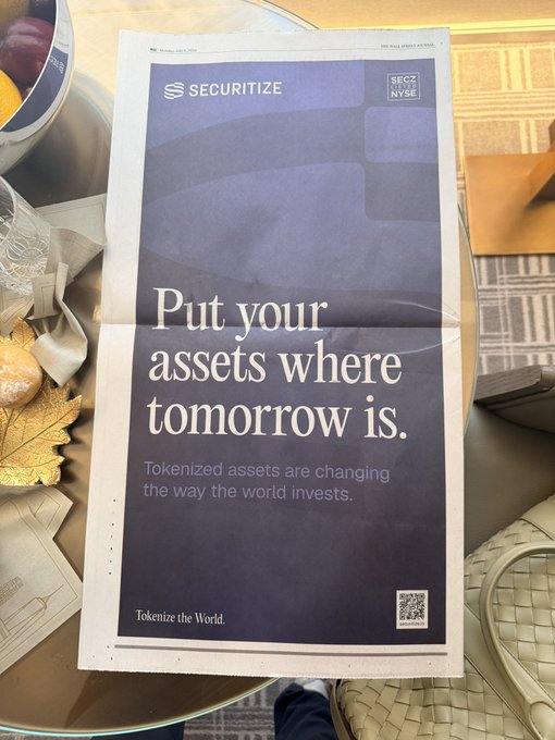 A Securitize newspaper advertisement displayed on a glass table, featuring the tagline "Put your assets where tomorrow is" and "Tokenized assets are changing the way the world invests," along with the hashtag "Tokenize the World" and SECZ NYSE listing details.
