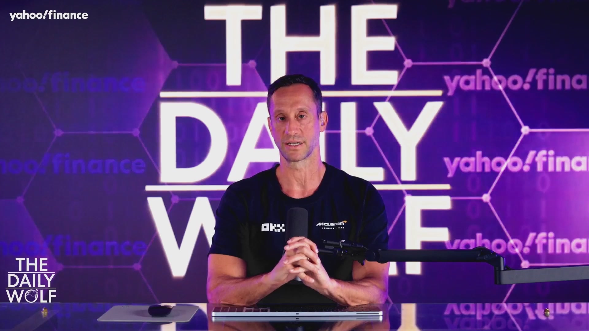 In the image, a man dressed in black is sitting behind a desk with his hands clasped in front of him as he appears to be talking into a microphone. The background features a purple color scheme that includes an abundance of white and purple text. One of the texts reads "LIVE NOW: Bitcoin Falls Below $68K As Outflows Accelerate!". This information is relevant to the image because it implies that someone might be discussing or reporting on the current state of Bitcoin, its value falling below $68,000, as well as the acceleration in outflow of Bitcoins. The context suggests that this event could be related to a financial news segment, possibly covering the effects of Bitcoin's price decrease and its increasing outflows.