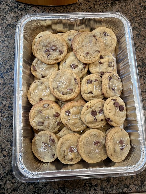 An aluminum catering tray is filled with approximately twenty freshly baked chocolate chip cookies sprinkled with flaky sea salt, sitting on a granite countertop.