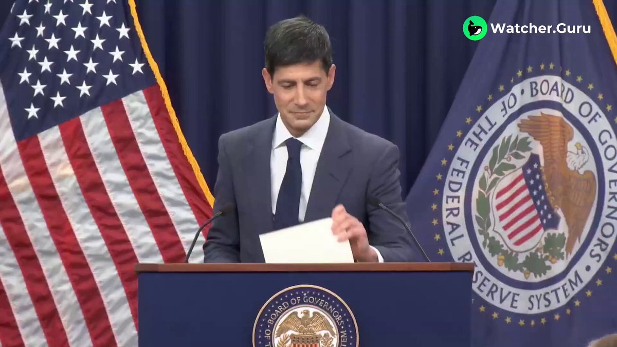 The image shows a man, possibly Kevin Warsh, standing behind a podium delivering a speech at a press conference. He is holding a piece of paper or a document as he speaks. The setting appears to be an official event with a backdrop featuring two American flags and a banner displaying "Watcher.Guru". The post also mentions that the Fed Chair rates are at an all-time high, indicating a significant economic development in real time.