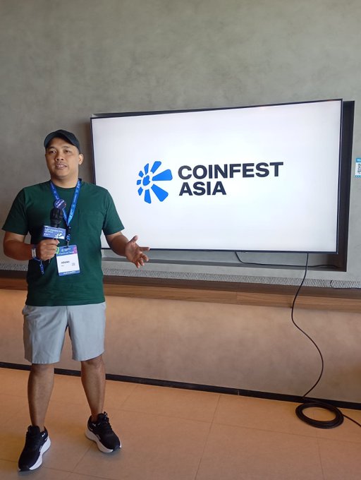 A man in a green shirt holds a microphone in front of a screen displaying the 'COINFEST ASIA' logo.