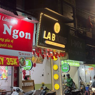 The image shows a street scene with a sign for "Lab Bong" hanging above a restaurant or food stand, likely indicating its name or location. A person can be seen riding a motorcycle past the establishment, while another individual is walking on the sidewalk nearby. The background includes other signs and buildings, suggesting that this area might be part of a larger commercial district.