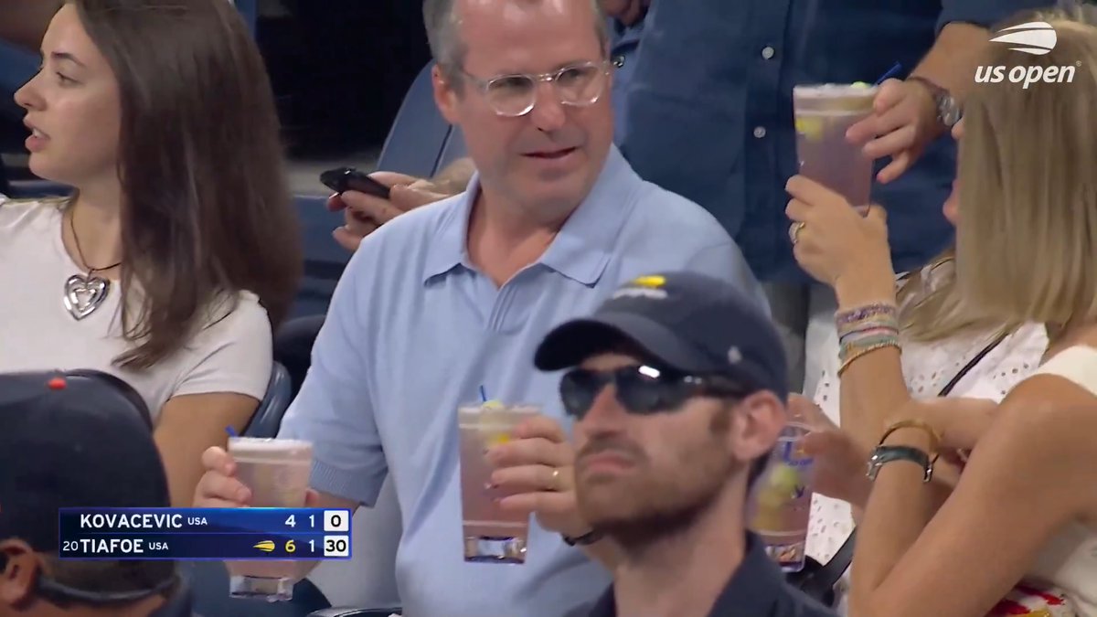 Spectators at the US Open hold clear cocktails resembling pickle juice drinks, watching a match where Tiafoe leads Kovacevic 6-4, 1-1 (30-0), with the man in sunglasses becoming the awkward center of attention.