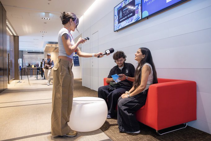 A woman with a face tattoo stands holding a microphone, interviewing two people seated on a red sofa in what appears to be a modern lobby or media area.