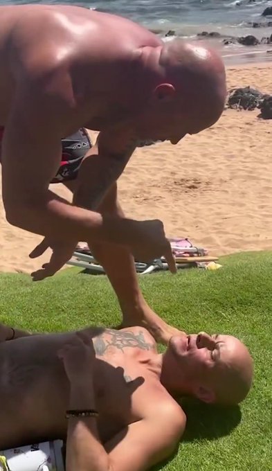 Two bald, muscular men outdoors on grass near a beach; one stands shirtless while the other lies on his back laughing as the standing man holds his arms down playfully.