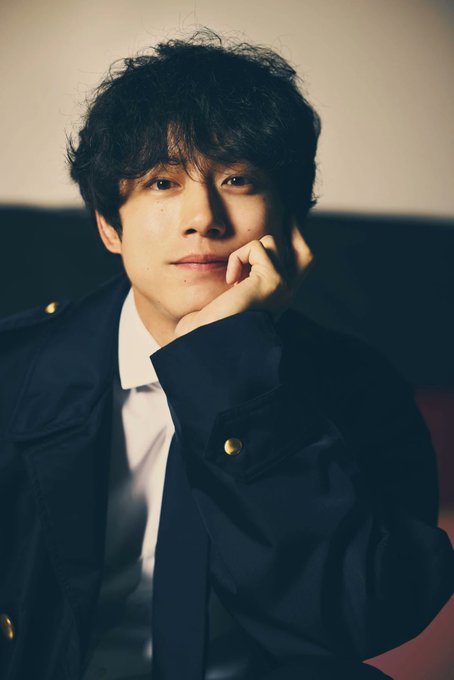 A portrait of a young man with dark, curly hair resting his chin on his hand, wearing a dark navy jacket with gold buttons over a white collared shirt.