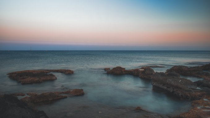 A serene seascape photograph captured at sunset or sunrise, showing rocks in the foreground with smooth, misty water, under a gradient sky transitioning from pink to blue.