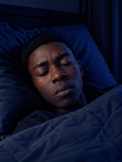 A close-up portrait of a young Black man sleeping peacefully in bed, wrapped in dark blue bedding under dim blue lighting.