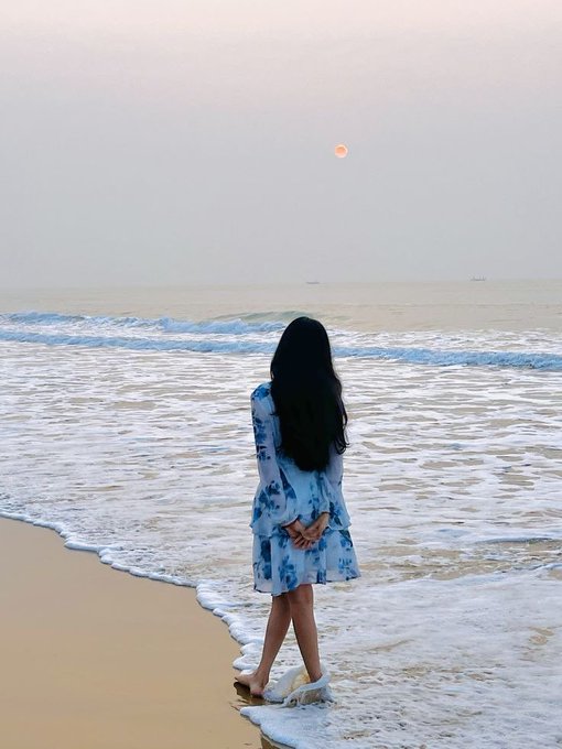 A photograph of a woman with long dark hair, seen from behind, wearing a light blue floral dress and standing barefoot at the edge of the ocean with waves washing over her feet.
