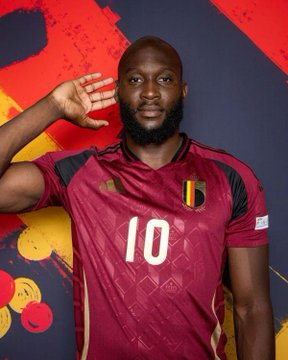 A man wearing a Belgium national team #10 jersey poses with a raised hand against a red backdrop.