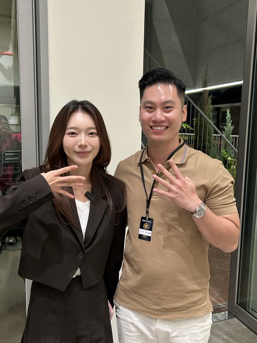 A woman in a black blazer and a man in a beige polo shirt wearing a lanyard pose together, both holding up peace signs and smiling.