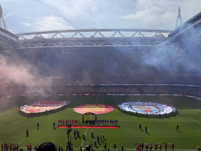 Two football teams, Arsenal and Manchester City, line up on the pitch at a packed stadium alongside large banners reading "Arsenal" and "Manchester City" with a central "Thank You" message, capturing the pre-match ceremony on a day out at the OKX-sponsored venue.