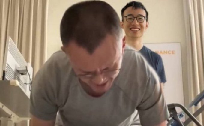 Two men in a room, with the foreground man in a gray shirt bending over laughing while a younger man with glasses smiles behind him; a framed Binance poster is visible in the background.