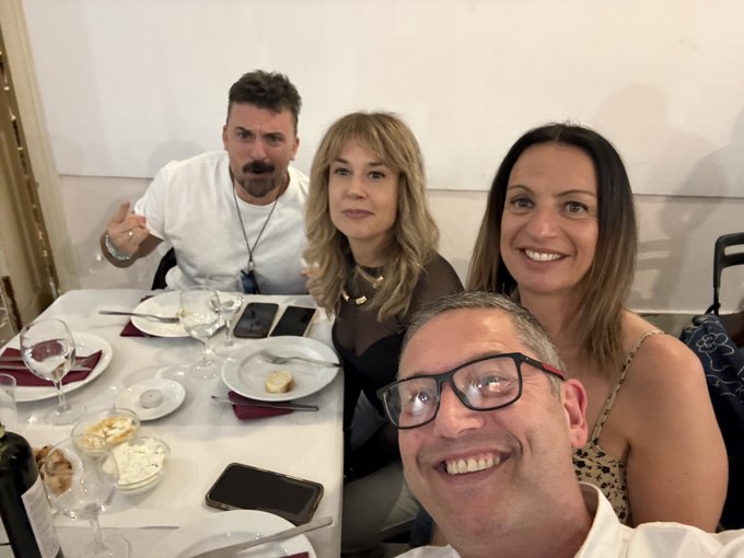 The image shows a group of four people sitting around a dining table, enjoying each other's company at an event. They are all smiling for the camera while posing for a picture. The table has various items such as wine glasses, cups, forks, knives, and bowls placed on it. There is also a cell phone visible in the scene. The background of the image features a wall with a painting hanging above the group, adding to the ambiance of the gathering.