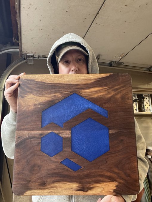 A person wearing a gray hoodie and a baseball cap holds up a square wooden plaque featuring a carved and blue-painted geometric logo.