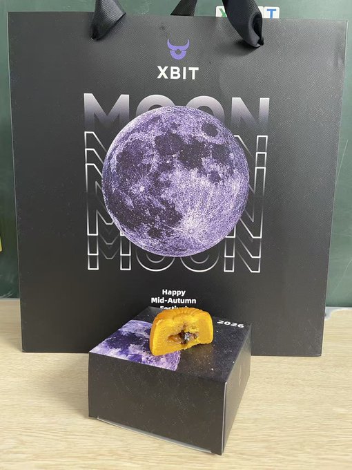 A black XBIT gift bag featuring a purple moon design with 'MOON MOON MOON' text and 'Happy Mid-Autumn Festival 2026' message, displayed next to a black box containing a cut mooncake.