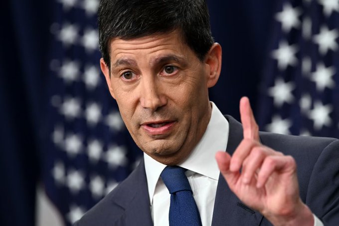 A close-up photograph of a man in a dark suit and blue tie speaking with his finger raised, set against a backdrop of American flags.