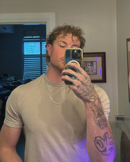 A muscular man with curly hair takes a mirror selfie while holding a smartphone, showing off heavily tattooed arms and wearing a tight beige t-shirt and silver chain.