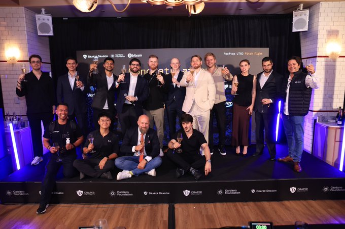 A group of people pose together holding champagne glasses on a stage with 'ReefTap UTXO' and 'Draper Dragon' branding, celebrating an event.