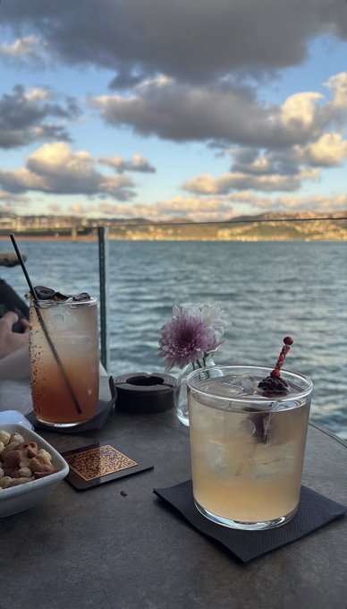 Two cocktails sit on a table by the water, with a tall dark drink garnished with cherries and a shorter amber drink garnished with a cherry, alongside a bowl of nuts and a pink flower, with a scenic bay and bridge visible in the background.