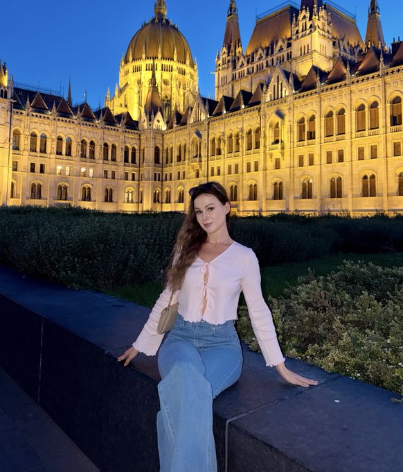 A young woman with long brown hair sits on a stone ledge wearing a white blouse and blue jeans, with the illuminated Hungarian Parliament Building in Budapest glowing in the background at dusk.