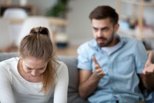 A man in a light blue shirt gestures while talking to a woman who appears upset, sitting on a gray couch.