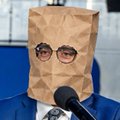 A person wearing a brown paper bag over their head with drawn-on glasses, sitting in front of a microphone against a blue background.