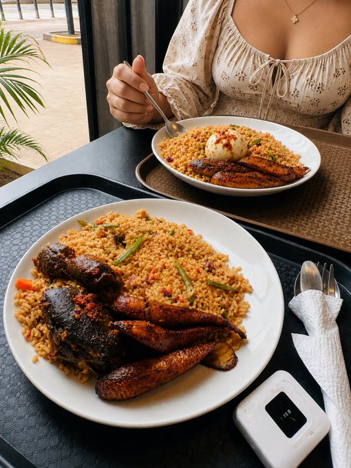 Two plates of food on a table, featuring rice, fried plantains, and meat dishes, with a person eating in the background and a portable power bank on the tray.