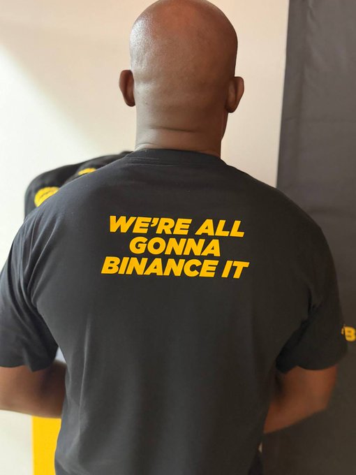 A man with a shaved head stands with his back to the camera, wearing a black t-shirt that reads "WE'RE ALL GONNA BINANCE IT" in bold yellow lettering—perfectly capturing the post's sentiment that the shirt's message speaks for itself.