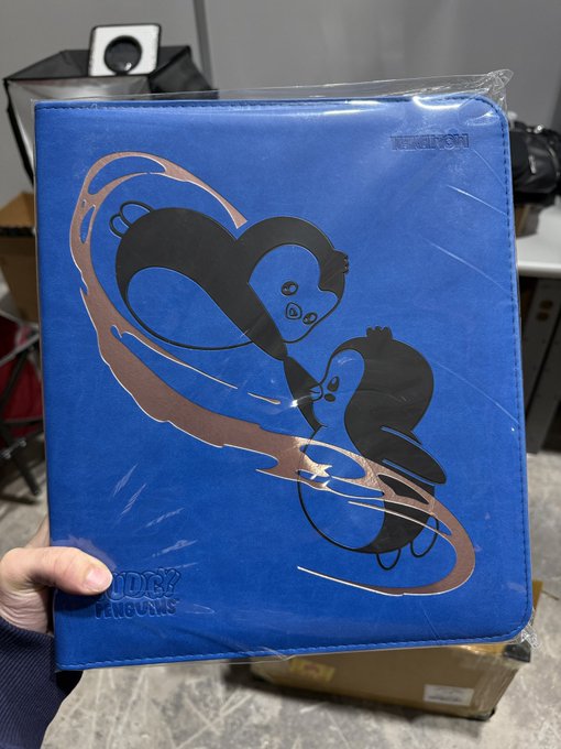 A person holds a blue Binder Albums portfolio still sealed in plastic, featuring an illustration of two black otters intertwined with a brown ribbon design on the cover.