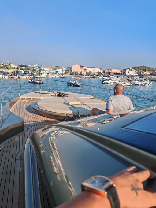 A first-person view from a yacht approaching a marina, showing a man's wrist with a watch in the foreground, another person in a white shirt seated ahead, and various boats and waterfront buildings in the distance.