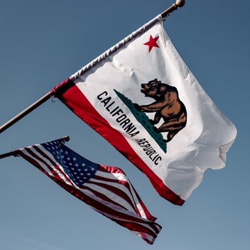The California Republic flag waves alongside the American flag against a clear blue sky, symbolizing the state's legislative action as its Senate passes a bill to ban platforms from listing memecoins issued by public officials after January 1, 2027.