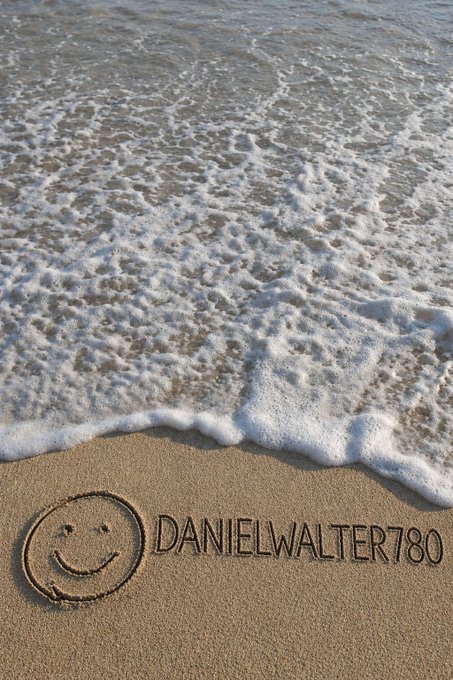 A smiley face and the text 'DANIELWALTER780' is drawn in the sand with foamy waves rolling in.