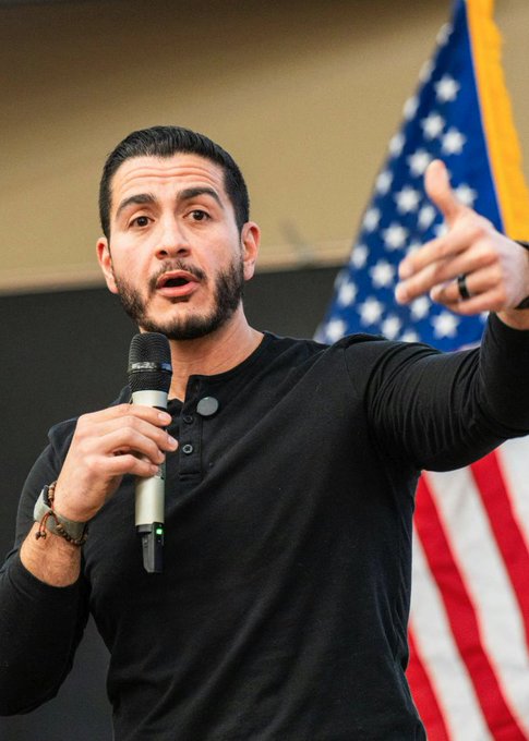A man with dark hair and a beard speaks passionately into a microphone while gesturing, with an American flag visible in the background. The image relates to the post about Abdul El-Sayed's projected primary victory in Michigan.
