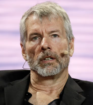 A close-up portrait of a middle-aged man with gray hair and a beard, wearing a headset microphone against a pinkish background.