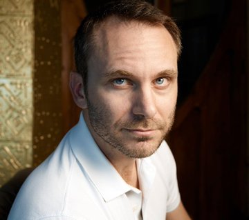 A photographic portrait of a man with short brown hair and light stubble, wearing a white polo shirt and looking directly at the camera.
