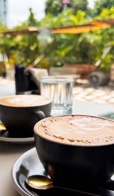 Two cups of coffee with frothy tops sit on a table at an outdoor café, with a glass of water and lush green plants in the background.