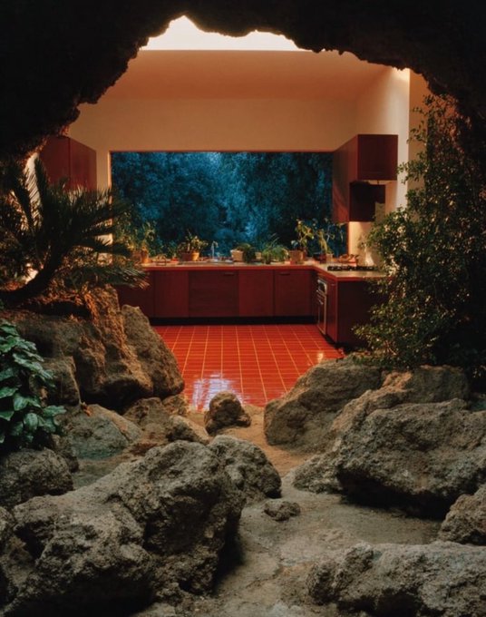 A view of a red-tiled kitchen interior seen through a rough rocky cave opening, with green plants and a dark windowed wall.