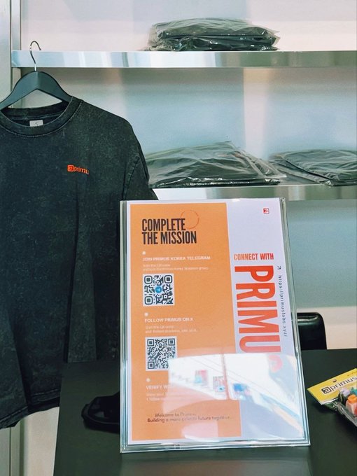 A retail display features a dark gray t-shirt with an orange 'primU' logo hanging next to a sign reading 'COMPLETE THE MISSION' with QR codes and instructions to connect with primU on Telegram and X, and verify attendance.