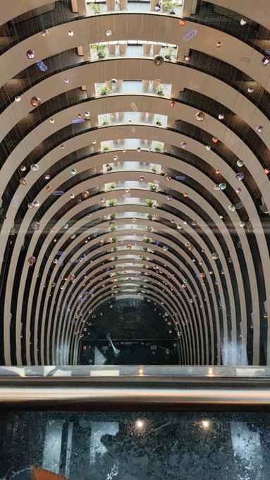 A dramatic upward view of a multi-story atrium with curved balconies and a decorative chandelier installation, looking up through an oculus at the top floors.